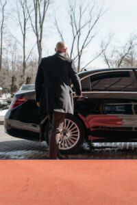 Bald man in a black suit approaches a luxury black car on a sunny day.